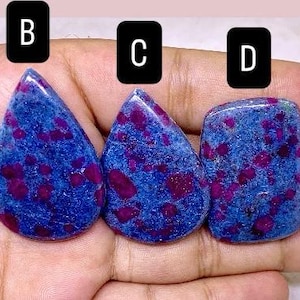 May include: Five polished gemstones with a blue base color and red inclusions. The stones vary in shape, including oval, teardrop, and rectangular. The gemstones are displayed against a light pink background.