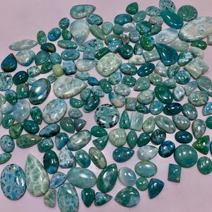 Puede incluir: Una colección de piedras preciosas pulidas de diversas formas y tamaños, que muestran una gama de tonos azules y verdes. Las piedras están dispersas sobre una superficie rosa claro, algunas con patrones y texturas únicos. El surtido incluye formas ovaladas, de lágrima y rectangulares.