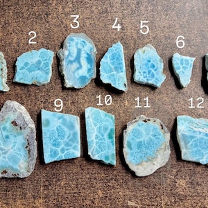 May include: A collection of twelve raw larimar gemstone pieces, each with unique blue and white patterns. The stones vary in shape and size, with a rough, natural texture. The gemstones are displayed on a brown surface.
