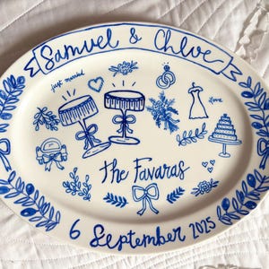 May include: Oval ceramic platter with a hand-painted blue design. The platter features the names "Samuel & Chloe" and "The Favaras", along with wedding-themed illustrations and the date "6 September 2025". Decorative floral and bow motifs frame the design.