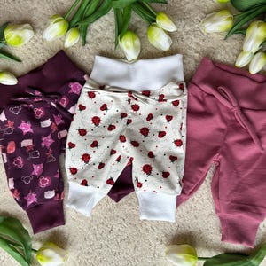 Könnte beinhalten: Drei Babyhosen in verschiedenen Farben und Mustern. Ein Paar ist weiß mit roten Marienkäfern, ein anderes ist tiefviolett mit einem rosa Muster, und das dritte ist einfarbig mauve. Alle haben elastische Bündchen und Manschetten.