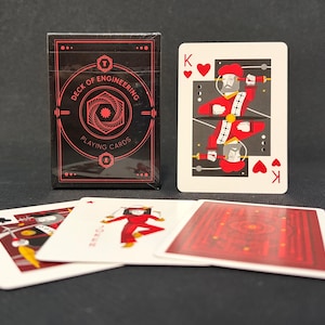 Puede incluir: Una caja negra etiquetada como Deck of Engineering Playing Cards con gráficos geométricos rojos, acompañada de un rey de corazones, un rey de tréboles, un comodín y un reverso de carta con un complejo patrón geométrico rojo y negro.