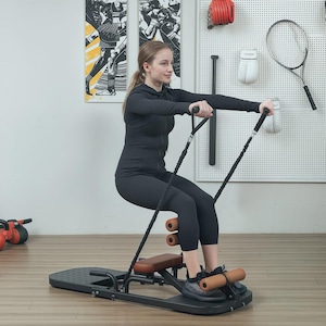 May include: A woman using a black and brown exercise machine. The machine features a black base, brown seat and leg supports, and black resistance bands. The woman is dressed in black athletic wear. The background includes sports-themed wall art and equipment.