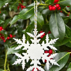 Op de afbeelding: Een witte sneeuwvlokversiering met ingewikkelde details hangt aan een goud-witte draad. De versiering staat tegen een achtergrond van groene bladeren en rode bessen, wat een feestelijke vakantiescène creëert.