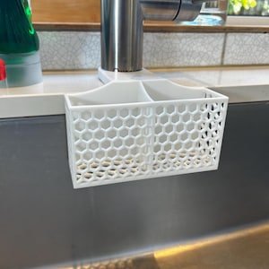 Può includere: Un organizer per lavello in plastica bianca con un design a nido d'ape. L'organizer ha due scomparti e si appende a un rubinetto. A sinistra dell'organizer è visibile una bottiglia verde di detersivo per piatti.