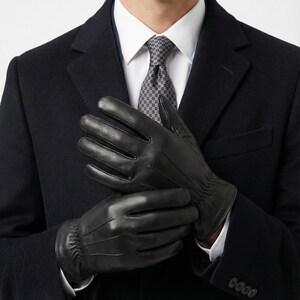 Pode incluir: Um par de luvas de couro preto com três linhas de costura decorativas no dorso da mão e um detalhe elástico franzido no pulso. As luvas são usadas com um casaco de lã azul-marinho, uma camisa branca e uma gravata cinza estampada.