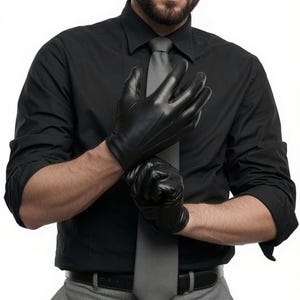 A man wearing a black shirt, grey tie, and grey trousers is pulling on black leather gloves.