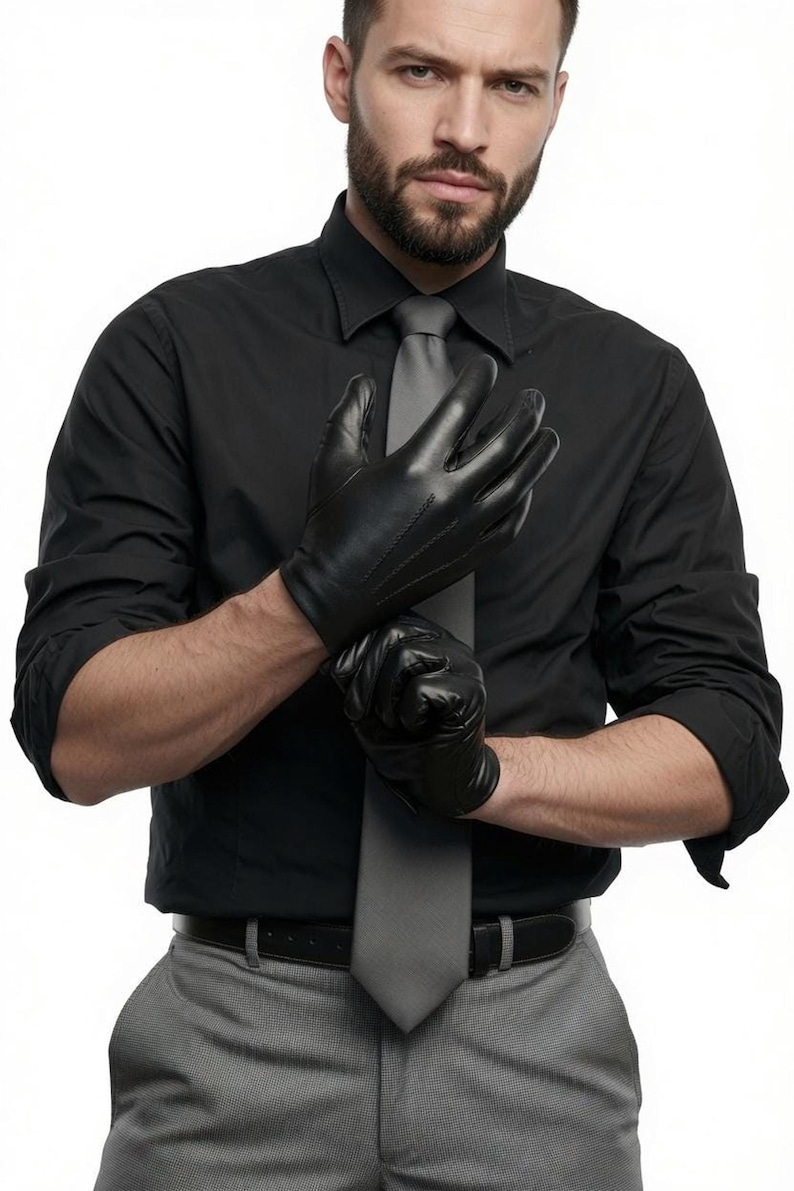 A man wearing a black shirt, grey tie, and grey trousers is pulling on black leather gloves.