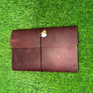 Può includere: Un diario in pelle bordeaux con chiusura a patta, assicurato da un elastico nero. Un piccolo ciondolo pende dalla fascia. Il diario è ambientato su uno sfondo di erba verde vibrante.