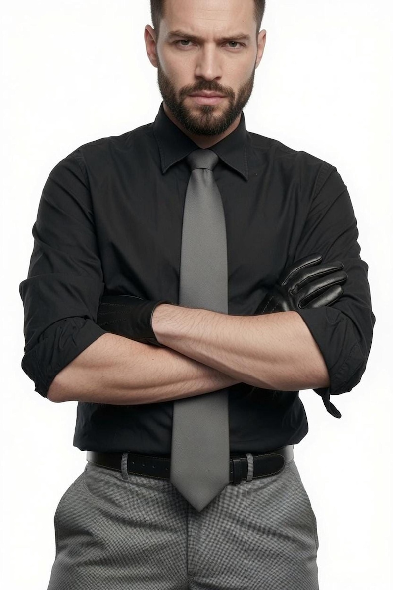 A man with crossed arms wearing a black shirt and grey tie wears black leather gloves.