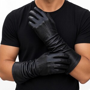 Pode incluir: Um par de luvas longas de couro preto com acabamento liso nos dedos e na mão, com detalhes franzidos que se estendem do pulso até o antebraço. As luvas apresentam três linhas de costura verticais no dorso da mão.