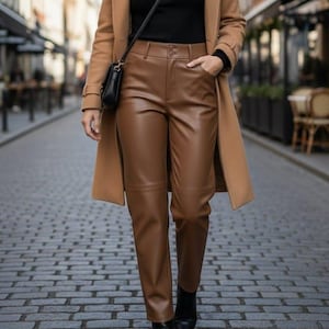 Op de afbeelding: Een outfit met een bruine leren broek, een zwarte top en een camelkleurige jas. De outfit wordt gecompleteerd met een zwarte tas en zwarte laarzen. De foto is genomen op een geplaveide straat.