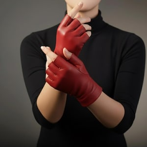 Op de afbeelding: Vingerloze rode leren handschoenen. De handschoenen zijn dieprood van kleur en hebben een gladde textuur. Het ontwerp heeft open vingers en een pasvorm tot aan de pols. De handschoenen worden gedragen met een zwarte top met lange mouwen.