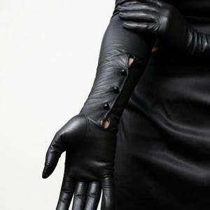 Könnte beinhalten: Ein Paar lange schwarze Lederhandschuhe, die über den Ellbogen reichen. Die Handschuhe haben eine eng anliegende Passform mit einer vertikalen Reihe von drei kleinen schwarzen Knöpfen am äußeren Unterarm, die eine Öffnung im Leder bilden.