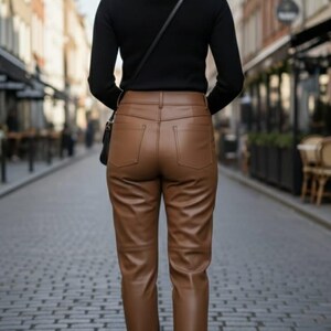 Op de afbeelding: Een persoon draagt een zwart shirt met lange mouwen en een bruine leren broek. De broek heeft een klassiek ontwerp met vijf zakken. De persoon draagt zwarte enkellaarsjes en een zwarte schoudertas. De achtergrond is een geplaveide straat.