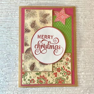 May include: A handmade Christmas card with a festive design. The card features a white circle with "MERRY Christmas" in red script, a red star, and patterned paper with pinecones and floral designs. The card is framed with pink and brown cardstock.
