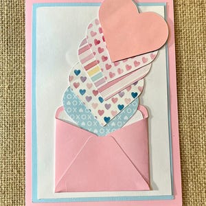 May include: A handmade greeting card with a pink envelope and layered heart cutouts. The hearts feature various patterns, including hearts, stripes, and XOXO designs. The card is framed with pink and light blue borders, creating a layered effect.