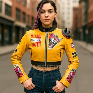 Può includere: Giacca corta in pelle gialla con finiture nere e chiusura a zip. La giacca presenta toppe con testo e disegni grafici, tra cui un disegno rosso e bianco sulla manica destra. È visibile un dettaglio nero sulla spalla.