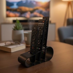 Könnte beinhalten: Ein schwarzer, geschwungener Fernbedienungshalter mit drei Fächern, der drei schwarze Fernbedienungen hält. Der Halter steht auf einem Holztisch, im Hintergrund ein Fernseher und eine Pflanze. Die Fernbedienungen haben verschiedene Knöpfe.