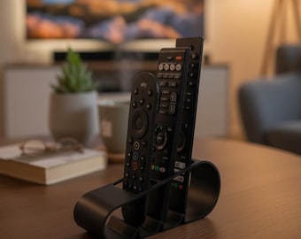 May include: A black, curved remote control holder with three compartments, holding three black remote controls. The holder sits on a wooden table, with a television and a plant in the background. The remote controls have various buttons.