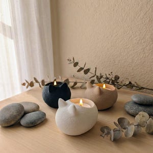 Könnte beinhalten: Drei katzenförmige Kerzen in Schwarz, Beige und Weiß, jede mit einem brennenden Docht. Die Kerzen stehen auf einer Holzoberfläche mit glatten Steinen und Zweigen. Der Hintergrund ist eine neutrale Wand und ein transparenter Vorhang.