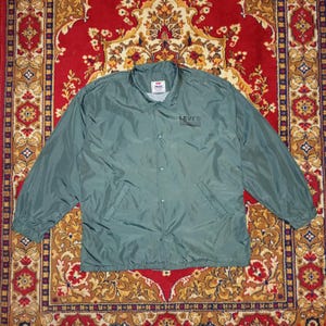 Puede incluir: Chaqueta de entrenador Levi's verde con cierre de botones, cuello y dos bolsillos delanteros. El logotipo de Levi's está impreso en la parte superior derecha del pecho. La chaqueta está tendida sobre una alfombra roja estampada.