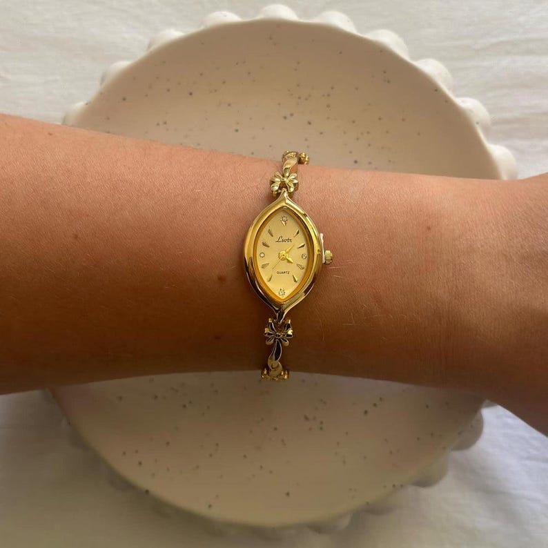 Op de afbeelding: Een goudkleurig quartz horloge met een ovale wijzerplaat en een delicate strikvormige band. De wijzerplaat is cr&egrave;mekleurig met gouden wijzers en uurmarkeringen. Het horloge wordt om de pols gedragen.