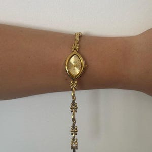 Op de afbeelding: Een goudkleurig polshorloge met een ovale wijzerplaat en een delicate bloemenarmband. De wijzerplaat van het horloge is goudkleurig met kleine diamantaccenten. De armband heeft een gedraaid ontwerp met bloemaccenten. Het horloge is ongeveer 18 cm lang.