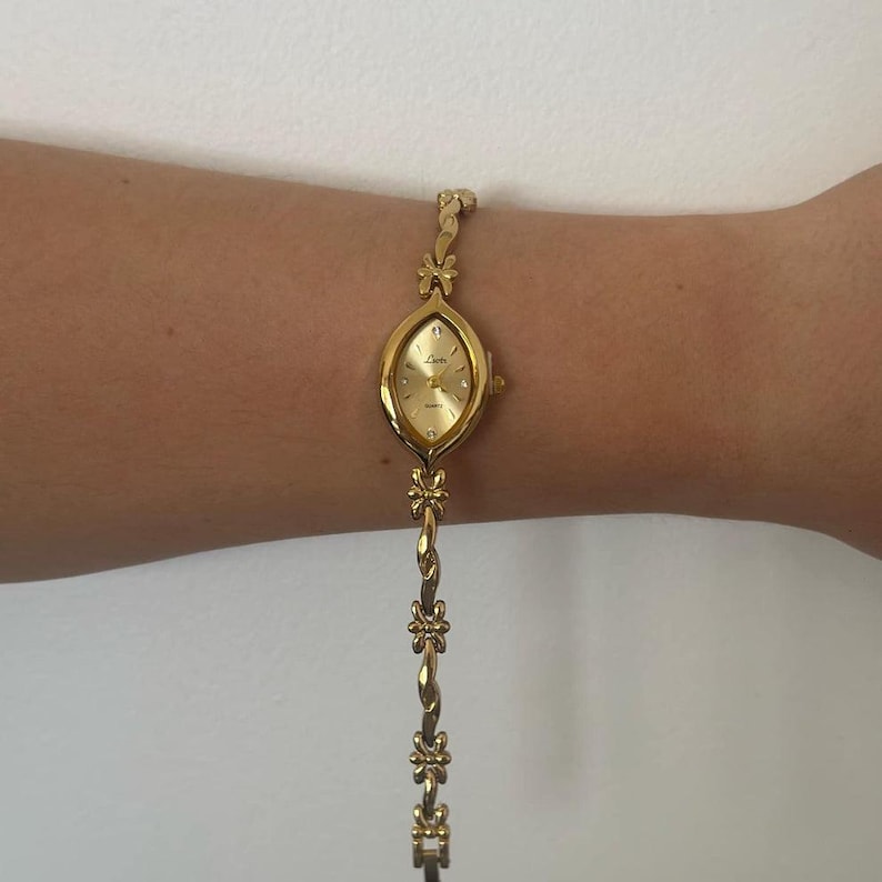Op de afbeelding: Een goudkleurig polshorloge met een ovale wijzerplaat en een delicate bloemenarmband. De wijzerplaat van het horloge is goudkleurig met kleine diamantaccenten. De armband heeft een gedraaid ontwerp met bloemaccenten. Het horloge is ongeveer 18 cm lang.