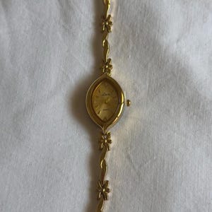 Op de afbeelding: Een goudkleurig polshorloge met een bloemmotief op de band. De wijzerplaat van het horloge is ovaalvormig met gouden wijzers en een gouden wijzerplaat. De band heeft kleine bloemaccenten. Het horloge staat op een witte achtergrond.