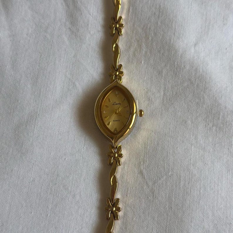 Op de afbeelding: Een goudkleurig polshorloge met een bloemmotief op de band. De wijzerplaat van het horloge is ovaalvormig met gouden wijzers en een gouden wijzerplaat. De band heeft kleine bloemaccenten. Het horloge staat op een witte achtergrond.