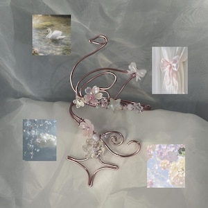May include: A rose-gold wire swan-shaped hair accessory, decorated with pink flowers, clear beads, and star-shaped details. The design includes a swan's head and neck, with a curved body. The background features a swan in water and other floral elements.