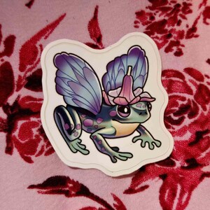 May include: A sticker of a frog with fairy wings and a flower crown. The frog is green with purple spots, and the wings are blue and purple. The background is pink with a floral pattern.