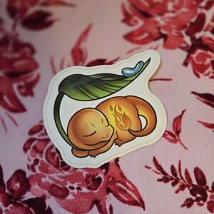 Puede incluir: Una pegatina con un personaje de dibujos animados de Charmander durmiendo bajo una hoja. El Charmander es naranja con una llama en la cola. La hoja es verde y una gota de agua azul está encima. La pegatina está sobre un fondo floral rosa.