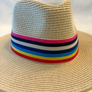 May include: A beige fedora-style hat with a wide brim. The hat features a colorful band with stripes of red, yellow, blue, navy, beige, pink, and green. The hat is made of woven material.
