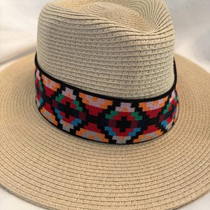 May include: A beige straw hat with a wide brim and a colorful patterned band. The band features a geometric design with red, blue, green, and orange accents. The hat has a classic fedora shape.