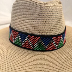 May include: A beige straw fedora hat with a black band featuring a colorful geometric pattern in red, green, blue, and white. The hat has a wide brim and a woven texture.