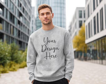 May include: A light grey sweatshirt with the text "Your Design Here" in a cursive font. The sweatshirt is long-sleeved and has a crew neck. The person is standing in front of a modern building.