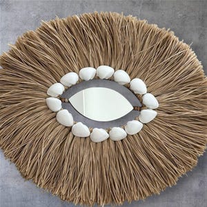 May include: Round wall mirror with a natural, woven raffia frame. The mirror is surrounded by white seashells and has a unique eye-shaped design. The neutral tones and coastal theme make it a decorative accent.