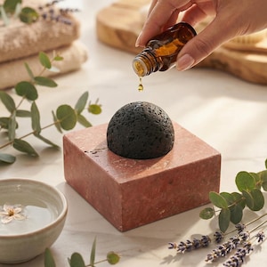 Pode incluir: Uma garrafa de vidro âmbar escuro despeja óleo sobre uma pedra preta e porosa. A pedra está sobre um bloco retangular avermelhado. Uma pequena tigela com água, uma flor, eucalipto e raminhos de lavanda completam a cena.