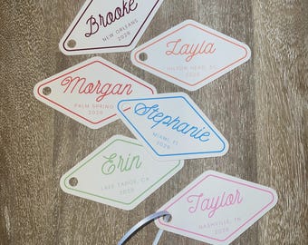 May include: A collection of diamond-shaped tags with names and locations printed on them. Each tag has a different color border and text color, including Brooke, Layla, Morgan, Stephanie, Erin, and Taylor, with the year 2026.