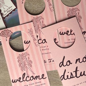 May include: Pink door hangers with vertical stripes and a circular cutout. The design includes illustrations of a showgirl, champagne bottles, and fans. Text reads "Welcome" and "Do Not Disturb" for a bachelorette party in New Orleans.