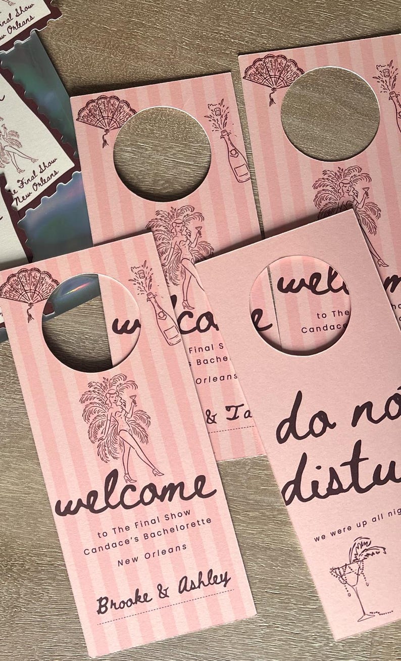 May include: Pink door hangers with vertical stripes and a circular cutout. The design includes illustrations of a showgirl, champagne bottles, and fans. Text reads "Welcome" and "Do Not Disturb" for a bachelorette party in New Orleans.
