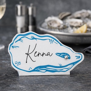 May include: A white oyster-shaped place card with a blue outline and the name "Kenna" in black script. A small silver bead is attached to the card. The card is standing on a dark gray surface.