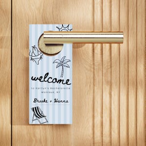 Może przedstawiać: Niebiesko-biały, pasiasty wieszak na drzwi z napisem "welcome" czarnym skryptem. Wieszak na drzwi ma motyw plażowy z ilustracjami bikini, palmy, słońca i krzesła plażowego. Tekst brzmi "to Kaitlyn's Bachelorette Montauk, NY" i "Brooke + Hanna."