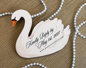 May include: A swan-shaped card with a white body, orange beak, and black accents. The text reads: Kindly Reply by May 1st, 2027, and WWW.WEDDINGWEBSITE.COM. The card is surrounded by a white beaded string, all set against a wood-grain surface.