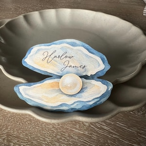Peut inclure: Une carte de place en forme de coquille d'huître, avec le nom "Harlow James" écrit en script élégant. La coquille est bleue et blanche, avec une perle au centre. La carte est posée sur une assiette grise.