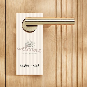May include: A cream and white striped door hanger with the word "welcome" and a building sketch. The text "to our wedding" and the names "hayley + nick" are also displayed. The hanger is on a wooden door with a gold-colored handle.