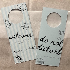 May include: Two light blue door hangers with a striped pattern. One says "welcome" with a palm tree and bikini graphic. The other says "do not disturb" with a cocktail graphic. Both have a circular cutout at the top.