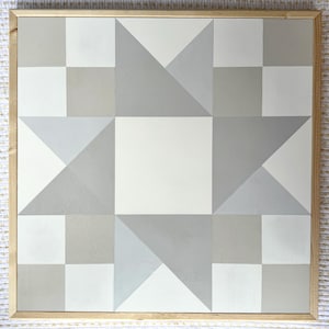 May include: Geometric wall art in a wooden frame. The design features a star pattern composed of gray, white, and light gray geometric shapes. The artwork is square and has a natural wood frame.